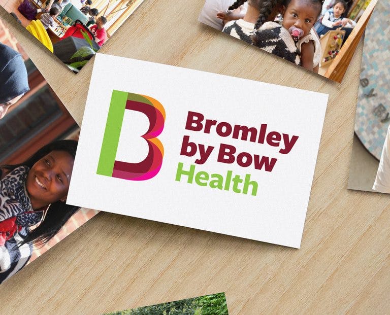 A postcard with the words Bromley by Bow Health on it is placed on a table. Next to it are two other pictures, one featuring a woman smiling, another featuring a baby being held by a girl. 